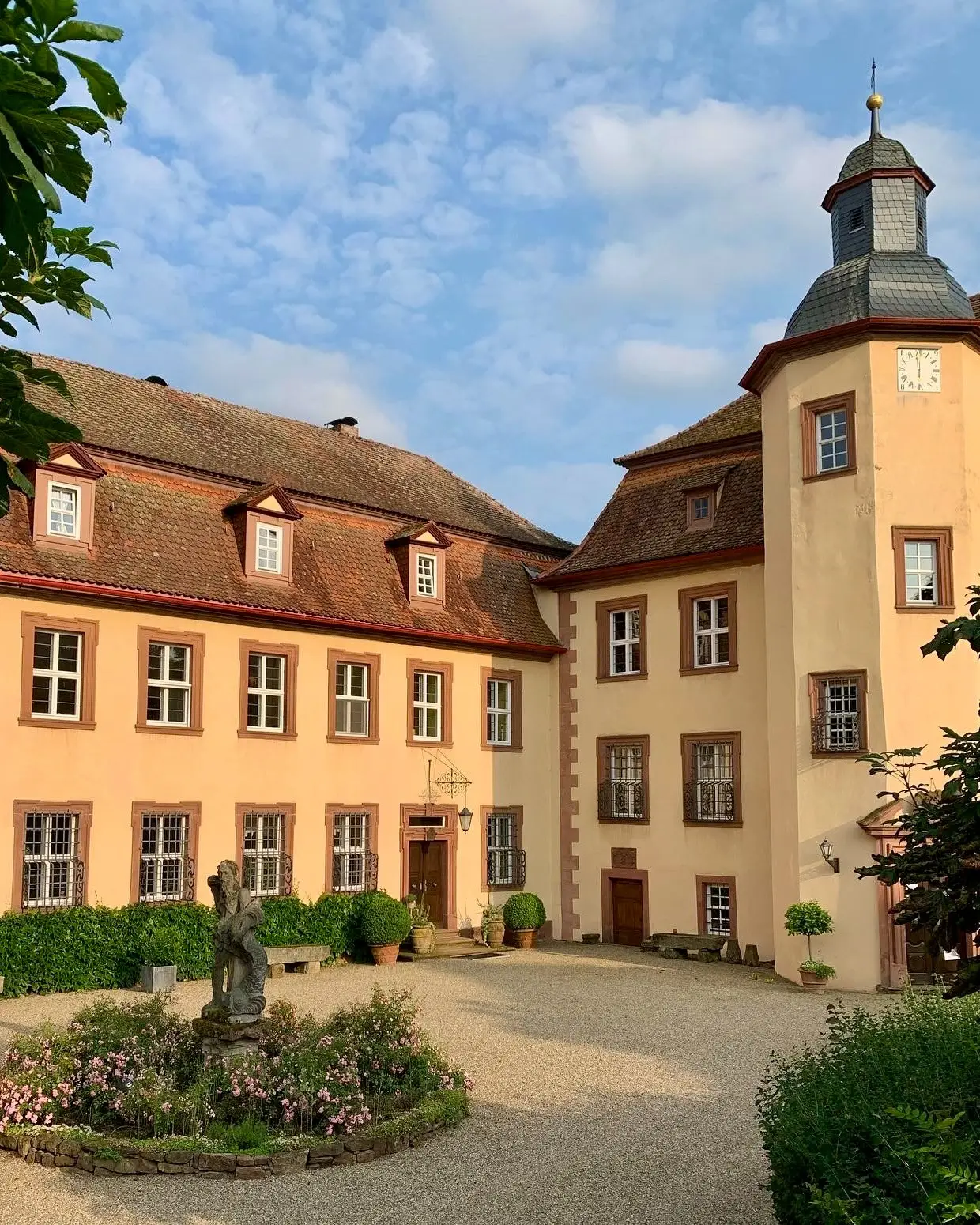 Innenhof von Schloss Roßbach mit apricotfarbener Fassade, Turm mit Laterne und einer Figur im Blumenbeet